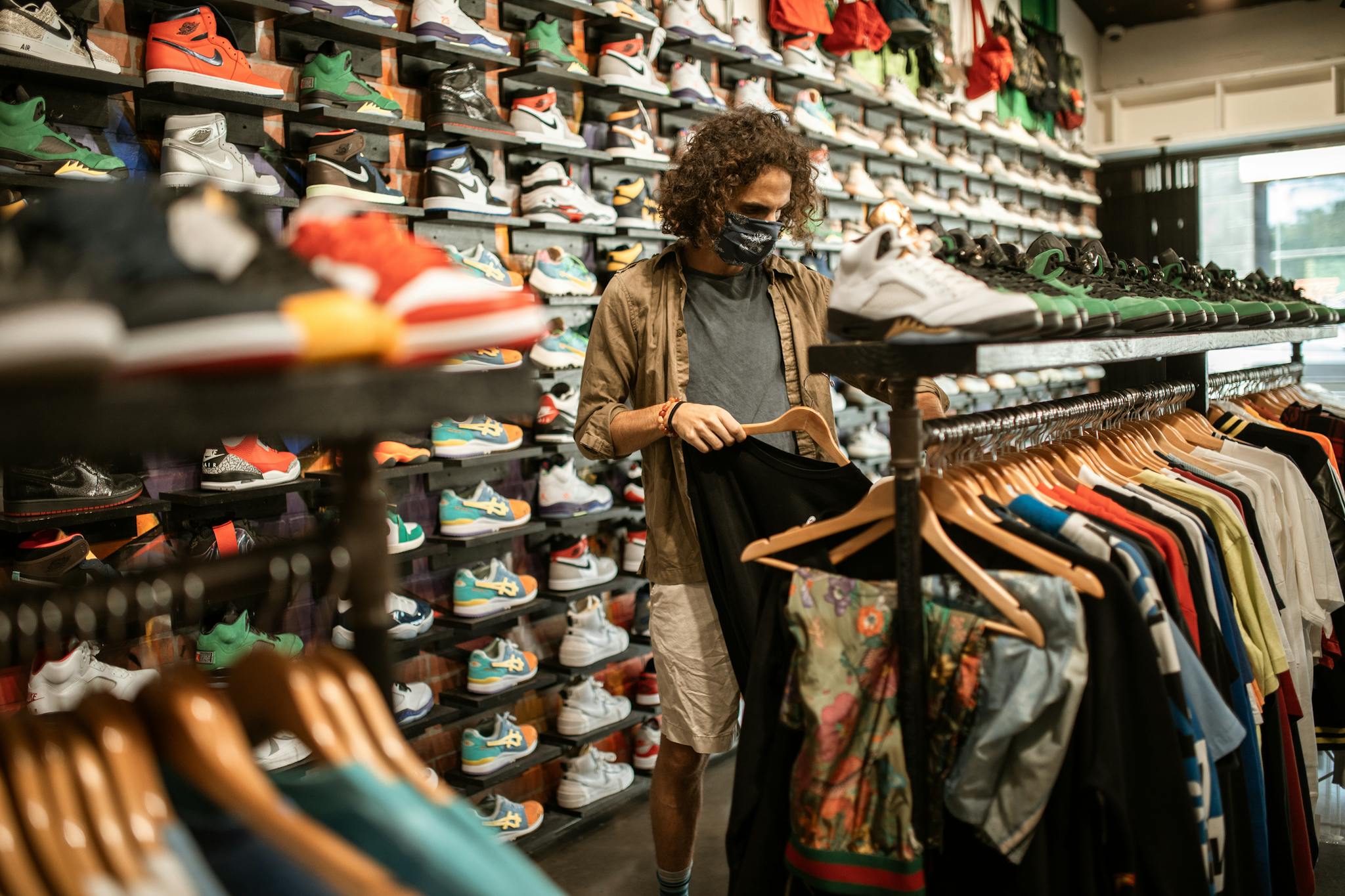 Young man in a sneaker and clothing store wearing a mask, browsing through shirts amid pandemic.
